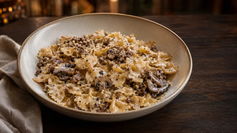 Eine Schüssel cremiger Pasta mit Hackfleisch und Pilzen, serviert auf einem dunklen Holztisch mit einer gefalteten Serviette.