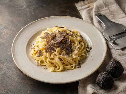 Tagliatelle mit schwarzen Trüffelspänen, ganzen Trüffeln und einem Trüffelhobel auf einer Stein-Arbeitsplatte.